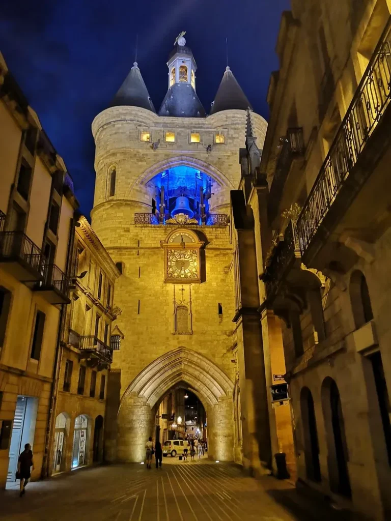 Un bâtiment historique en pierre avec une entrée voûtée et une tour d'horloge bleue illuminée la nuit.