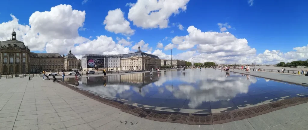 Vue panoramique d'une place de ville, avec une fontaine centrale, entourée de vie urbaine et de bâtiments historiques.