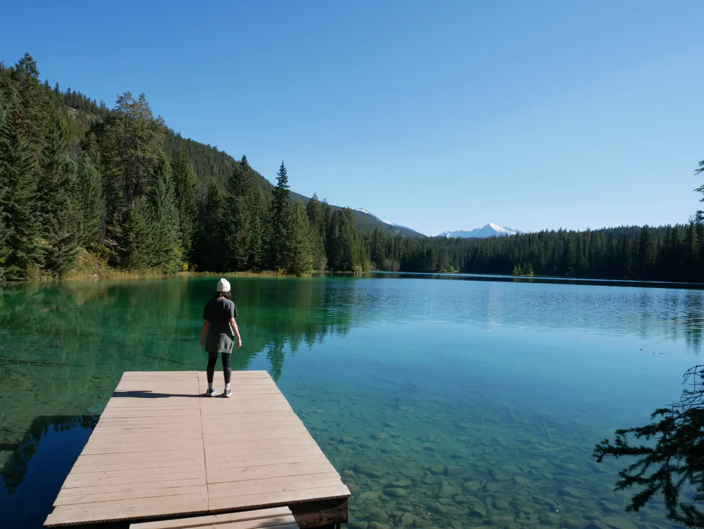 Gwen faisant face au lac turquoise entouré de sapins avec des montagnes en arrière plan